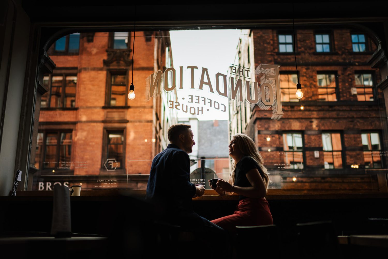 A young couple enjoying a coffee looking out of a window whilst on a Northern Quarter Engagement Session before their wedding